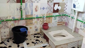 A bathroom with a toilet, a bucket, a red pot, and a shower area with a green pipe.