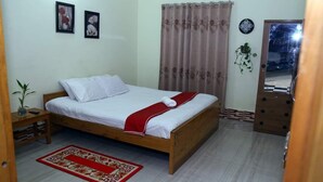 Un dormitorio con una cama de madera, ropa de cama blanca, una alfombra estampada en rojo y blanco, una mesita de noche de madera, una planta en maceta y una puerta con un espejo.