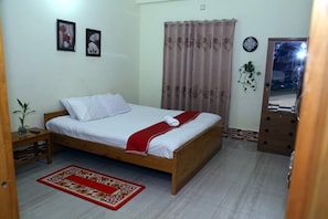A bedroom with a wooden bed, white bedding, a red and white patterned rug, a wooden nightstand, a potted plant, and a door with a mirror. - Brishti Bilash Guesthouse (Sreemangal)