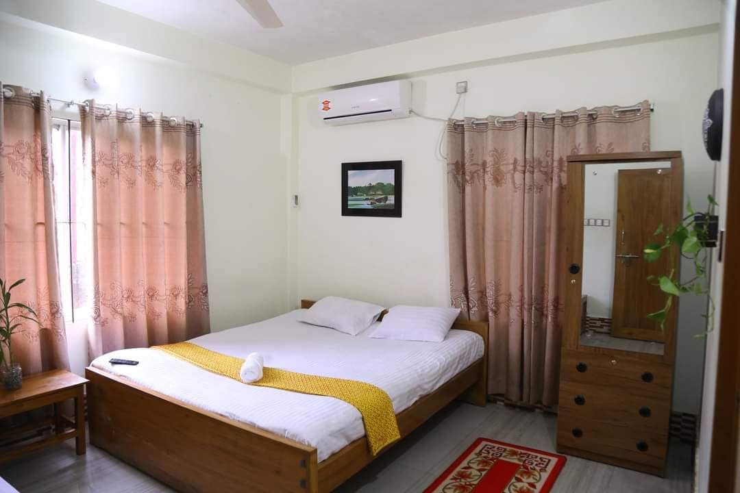 Een slaapkamer met een bed, nachtkastje, spiegel, raam met gordijnen, een airconditioning en een lijst met een afbeelding aan de muur.