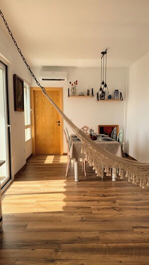 Een kamer met houten vloer, een eettafel met stoelen, een hangmat en een houten deur.