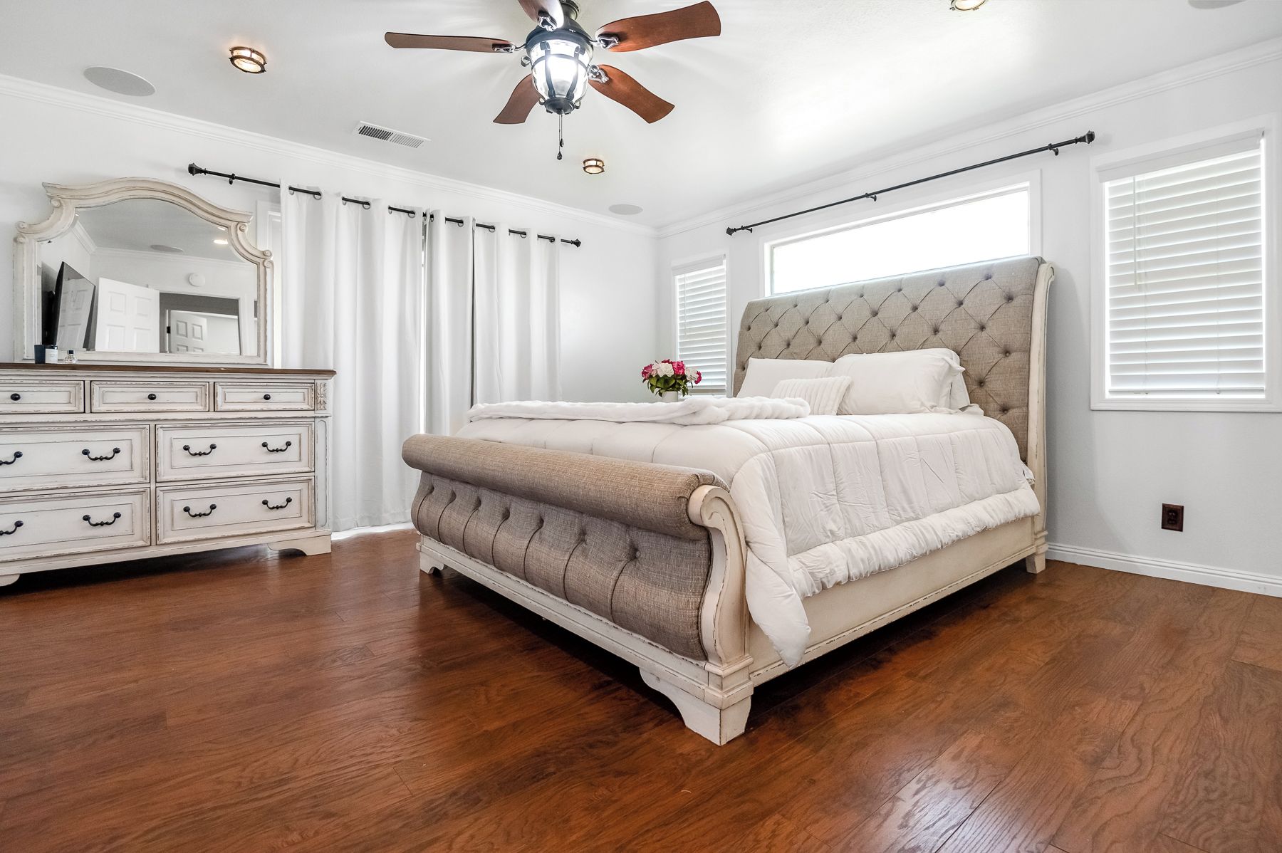 A bedroom with a large bed, a dresser, a ceiling fan, and a window with blinds.