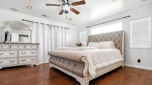 A bedroom with a large bed, a dresser, a ceiling fan, and a window with blinds. - Casa Luna, Modern Comfort (Visalia)