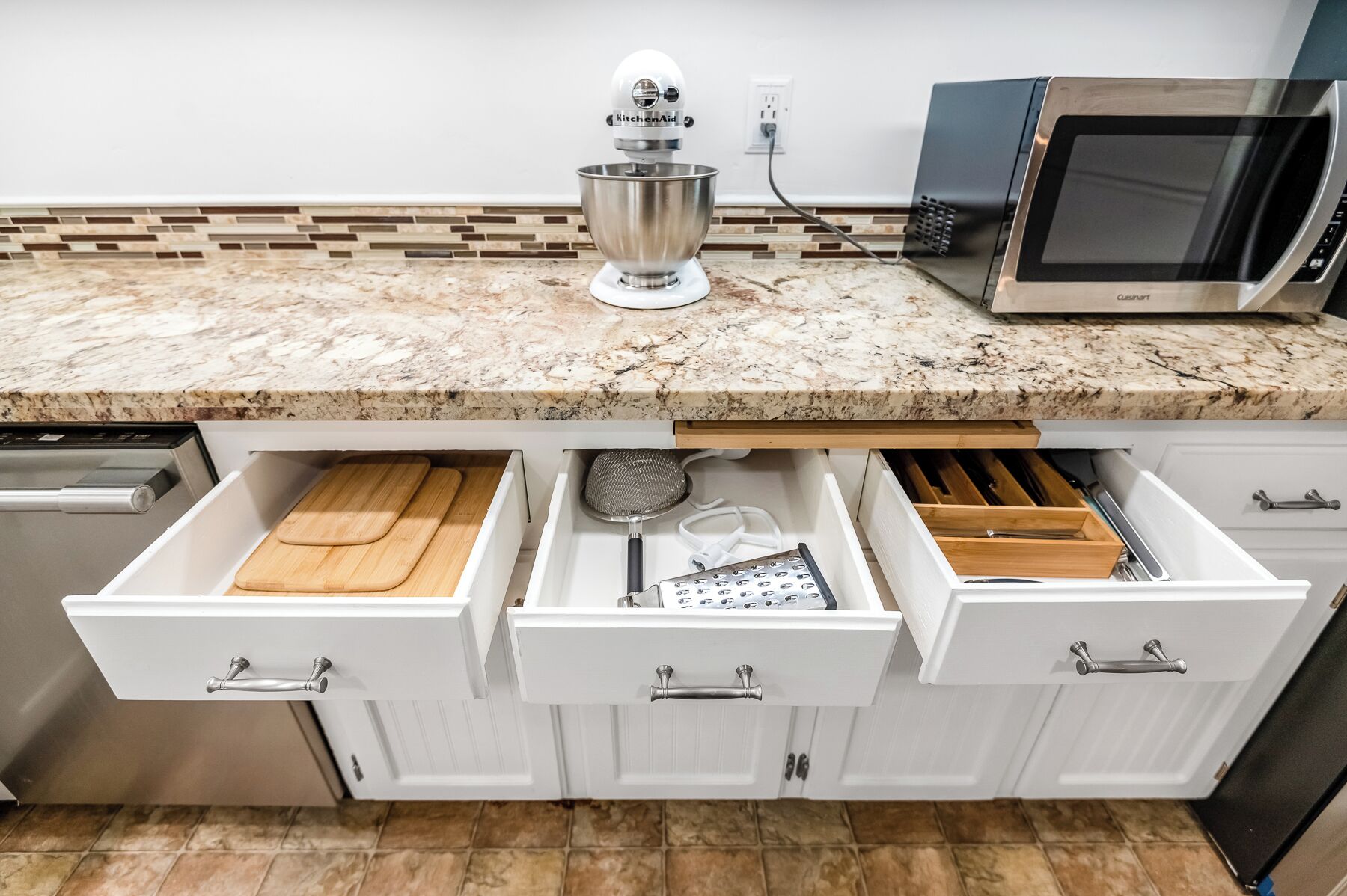 A kitchen with a granite countertop, a KitchenAid mixer, a microwave, and three drawers with kitchen utensils.