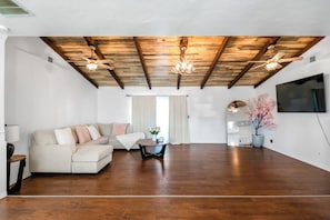 A spacious living room with a wooden ceiling, a sofa, a coffee table, and a TV mounted on the wall.