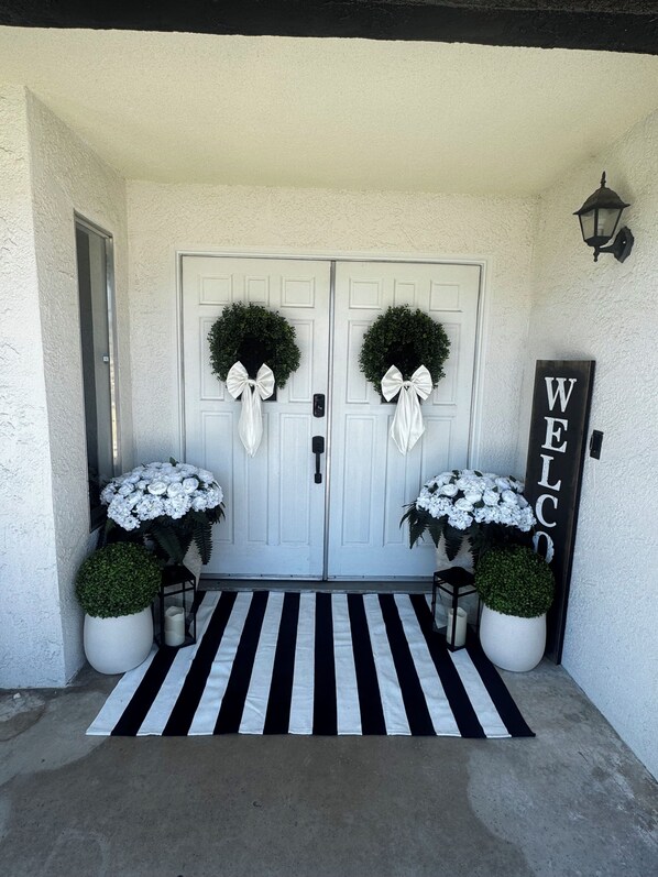 A double door with wreaths and a welcome sign, flanked by potted plants and a black-and-white striped mat.
