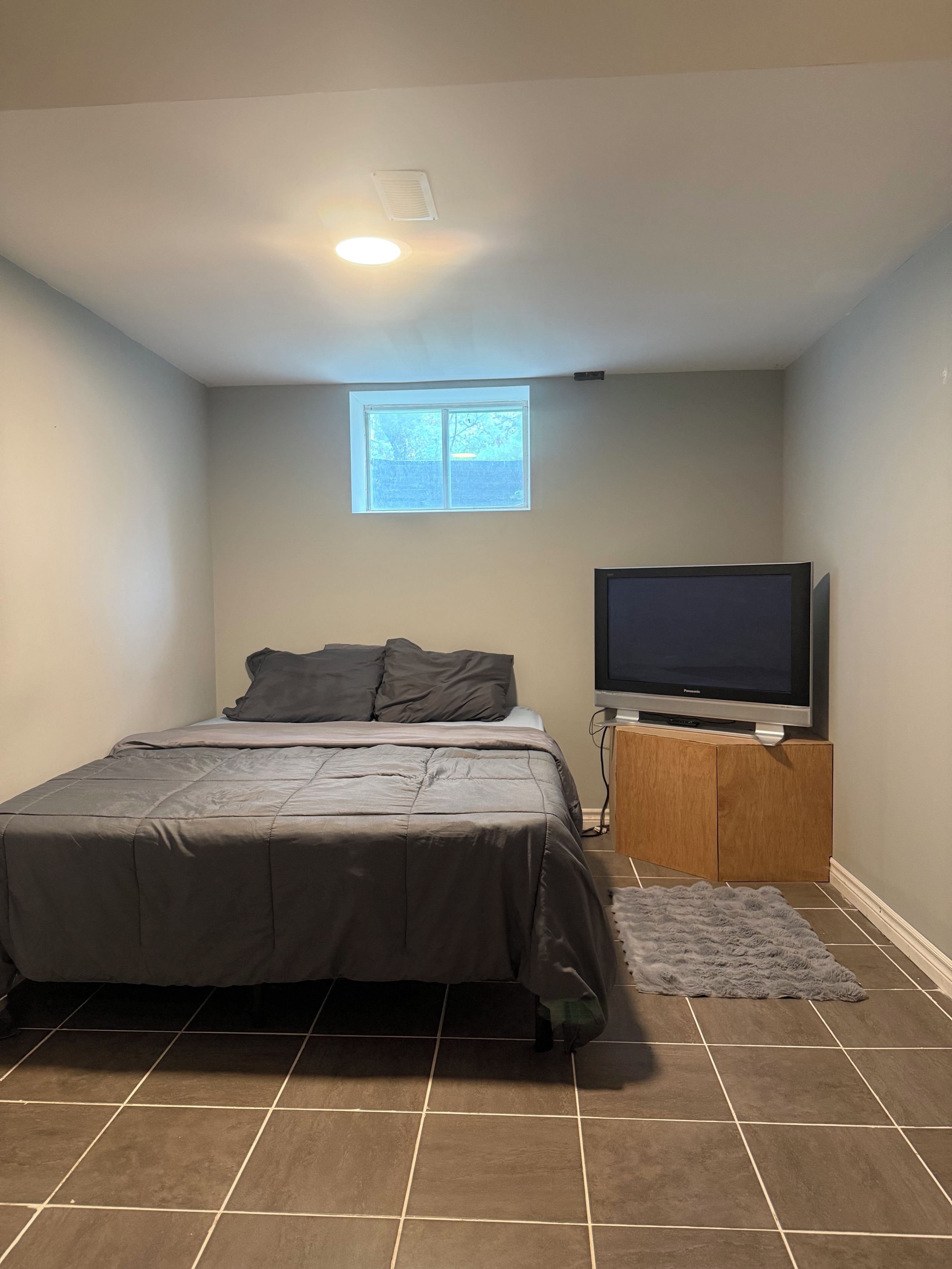 Une chambre à coucher avec un lit, un téléviseur posé sur un meuble en bois, une fenêtre et un luminaire au plafond.