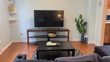 Een woonkamer met een flatscreen televisie op een standaard, een bijzettafeltje, een bank, een potplant en een klok aan de muur.
