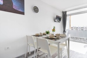 A dining area with a table set for two, white chairs, a clock on the wall, and a window with a view of buildings. - Blue sky:  ocean view & 5 min to Jumbo (Playa del Ingles)