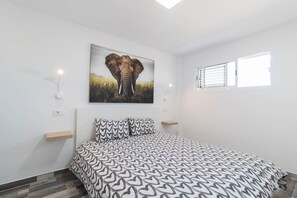 Ein Schlafzimmer mit einem Bett, einem an der Wand befestigten Elefantenbild, Nachttischen und einem Fenster mit Jalousien.