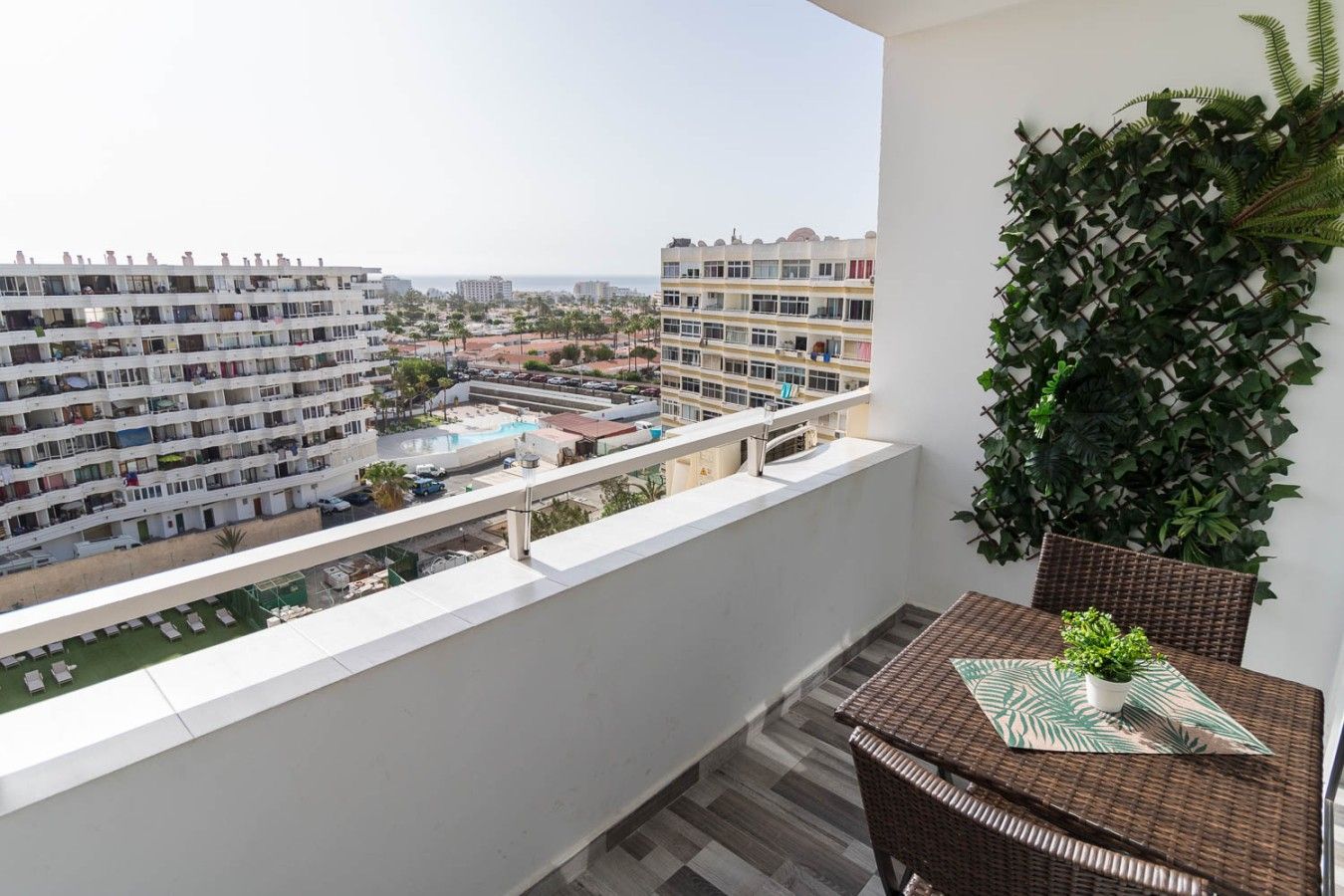 A balcony with a view of a residential area, a wicker table with a plant, and a green leafy wall.