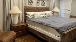 Un dormitorio con una cama grande, dos lámparas de noche, una mesita de noche de madera, una silla de mimbre, una puerta y un equipo de aire acondicionado en la pared.