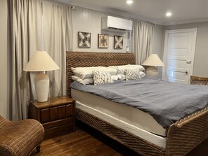 A bedroom with a large bed, two bedside lamps, a wooden nightstand, a wicker chair, a door, and an air conditioning unit on the wall. - Spacious 3-bedroom condo with pool and laundry, steps from beach and boardwalk! (Wildwood)