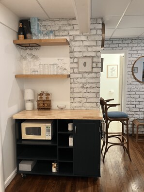 Ein Küchenbereich mit einer Holzausziehtafel, einer Mikrowelle und einem schwarzen Schrank. Darüber befinden sich Regale mit Gläsern und ein gerahmtes Bild an der Wand.