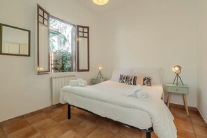 A bedroom with a bed, two bedside tables with lamps, a window with a view of greenery, and a radiator.