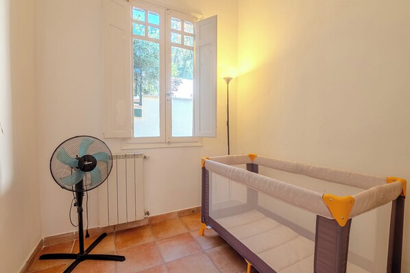 A room with a standing fan, a window with open shutters, and a bed with a safety rail.