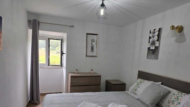 Ein Schlafzimmer mit einem Bett, einem Nachttisch, einer Kommode, einem Fenster mit Vorhängen und einer Wandleuchte.