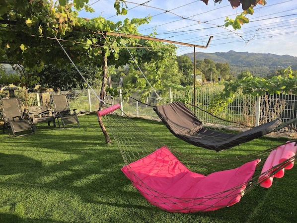 Una hamaca instalada en una zona con césped, una valla metálica y sillas, con vistas a un viñedo y colinas.