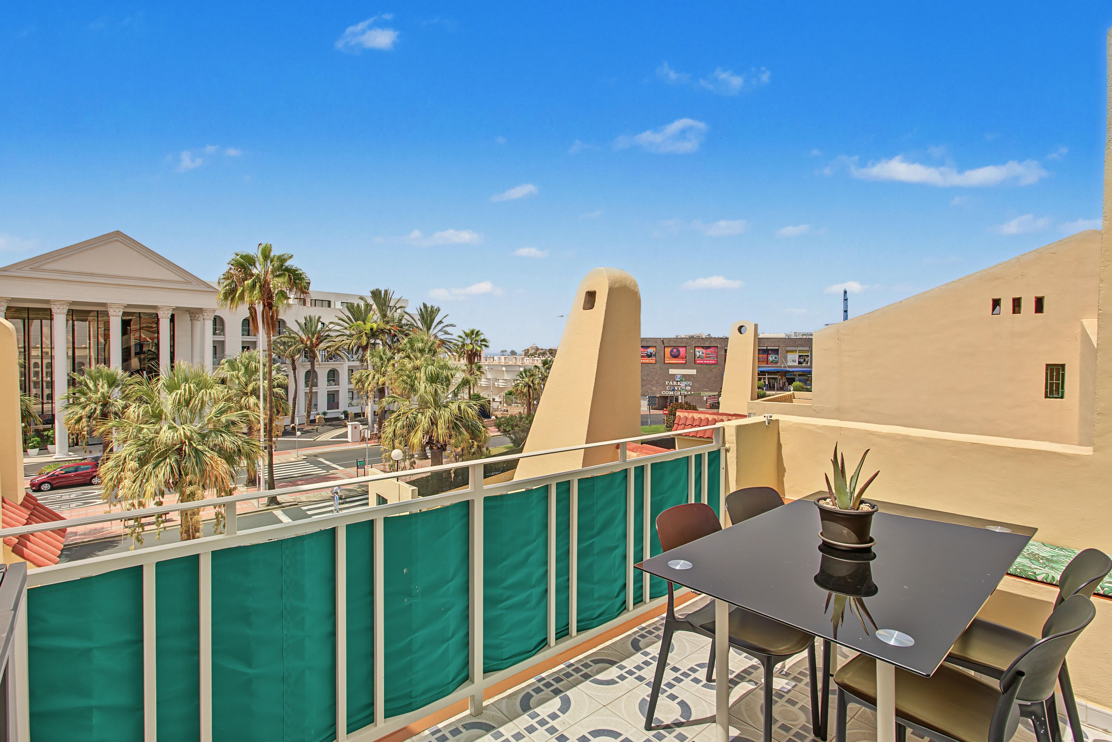 Un balcone con tavolo e sedie, una pianta in vaso e vista su un edificio con colonne e palme.