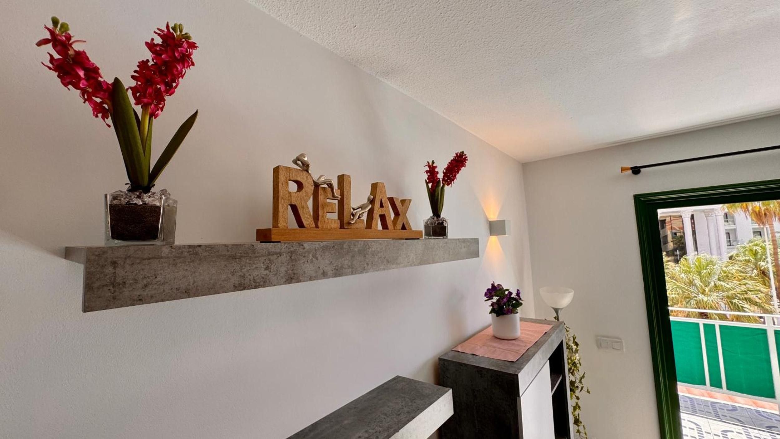 Een modern interieur met een plank waarop bloemen staan, een decoratief bord met de tekst 'RELAX' en een kleine tafel met een lamp.