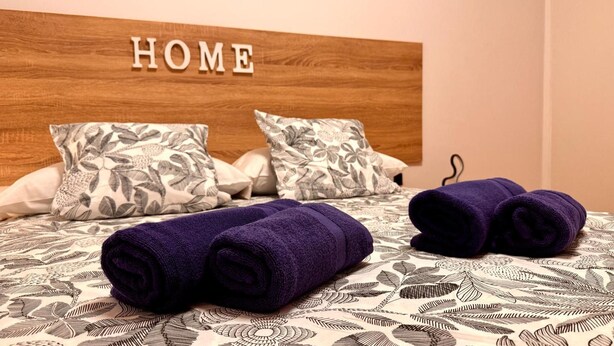 Una cama bien hecha con una colcha de motivos florales, dos toallas moradas enrolladas y un cabecero con la palabra 'HOME' escrita.