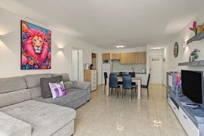 A modern living room with a grey sofa, a dining area with a wooden table and chairs, a kitchen with wooden cabinets, and a large TV on the right. - Spacious and Bright 'Mareverde' Apartment with Sea Views, Pool, and WiFi (Adeje)