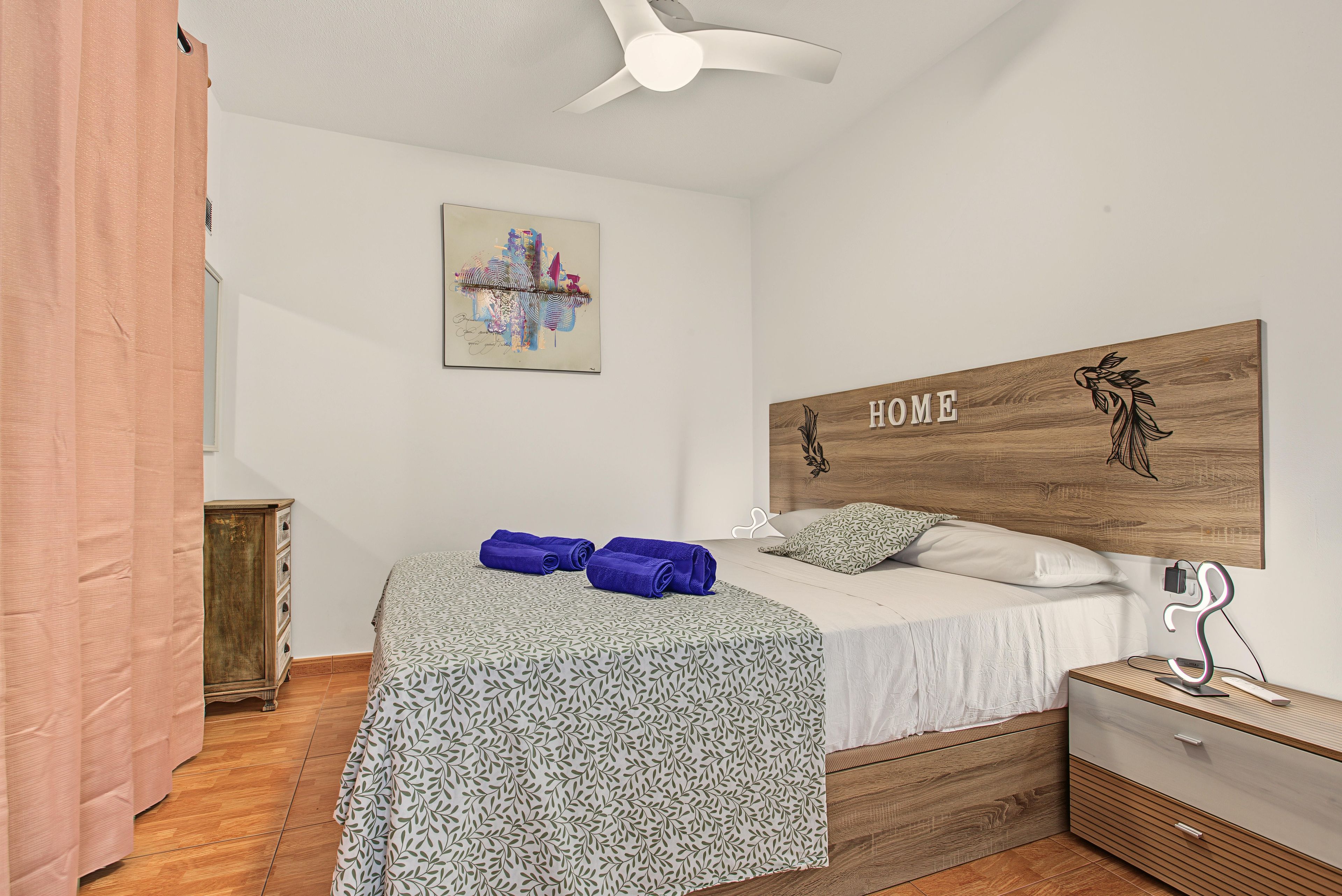 A bedroom with a bed, a headboard with 'HOME' written on it, a ceiling fan, a small wooden cabinet, and a wall art piece.