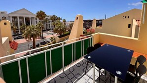 Un balcón con mesa y sillas, con vista a una calle con palmeras y edificios.