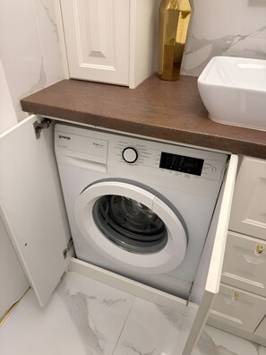 Lavadora compacta e integrada con panel de control, ubicada debajo de una encimera en un baño con suelo de mármol.