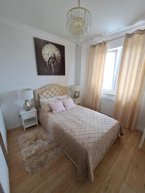 Un dormitorio con una cama tamaño reina, una mesita de noche, una lámpara, un candelabro, una ventana con cortinas y una obra de arte en la pared.