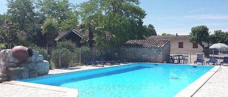 A swimming pool with lounge chairs, a stone wall, and a small building with a tiled roof.