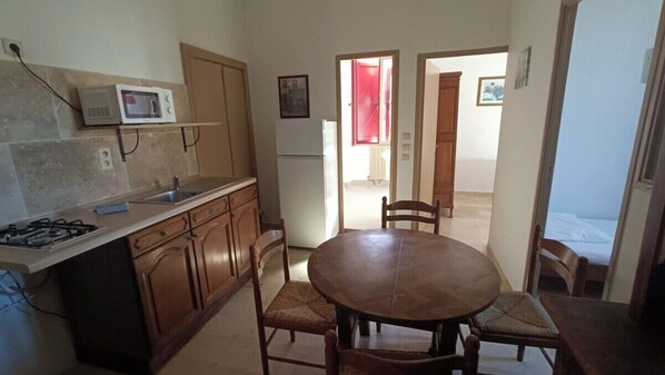 Une cuisine compacte équipée d’un four à micro-ondes, d’une cuisinière et d’un évier, un coin repas avec une table et des chaises, et une vue sur une chambre à coucher par une porte ouverte.