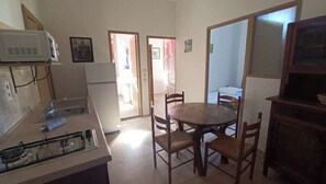 Une cuisine compacte équipée d’une cuisinière à gaz, d’un four à micro-ondes et d’un réfrigérateur. On y trouve une table à manger avec des chaises et un petit meuble de rangement.