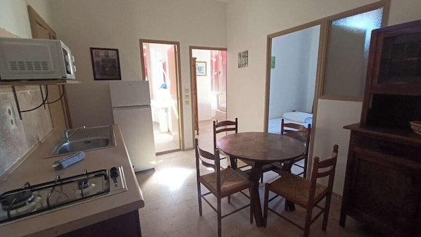 A compact kitchen with a gas stove, microwave, and refrigerator. There is a dining table with chairs and a small cabinet.