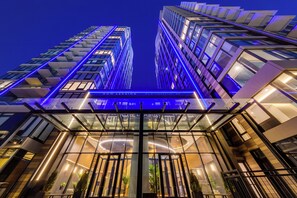 A modern hotel entrance with a glass facade, illuminated signage, and a view of the surrounding high-rise buildings.