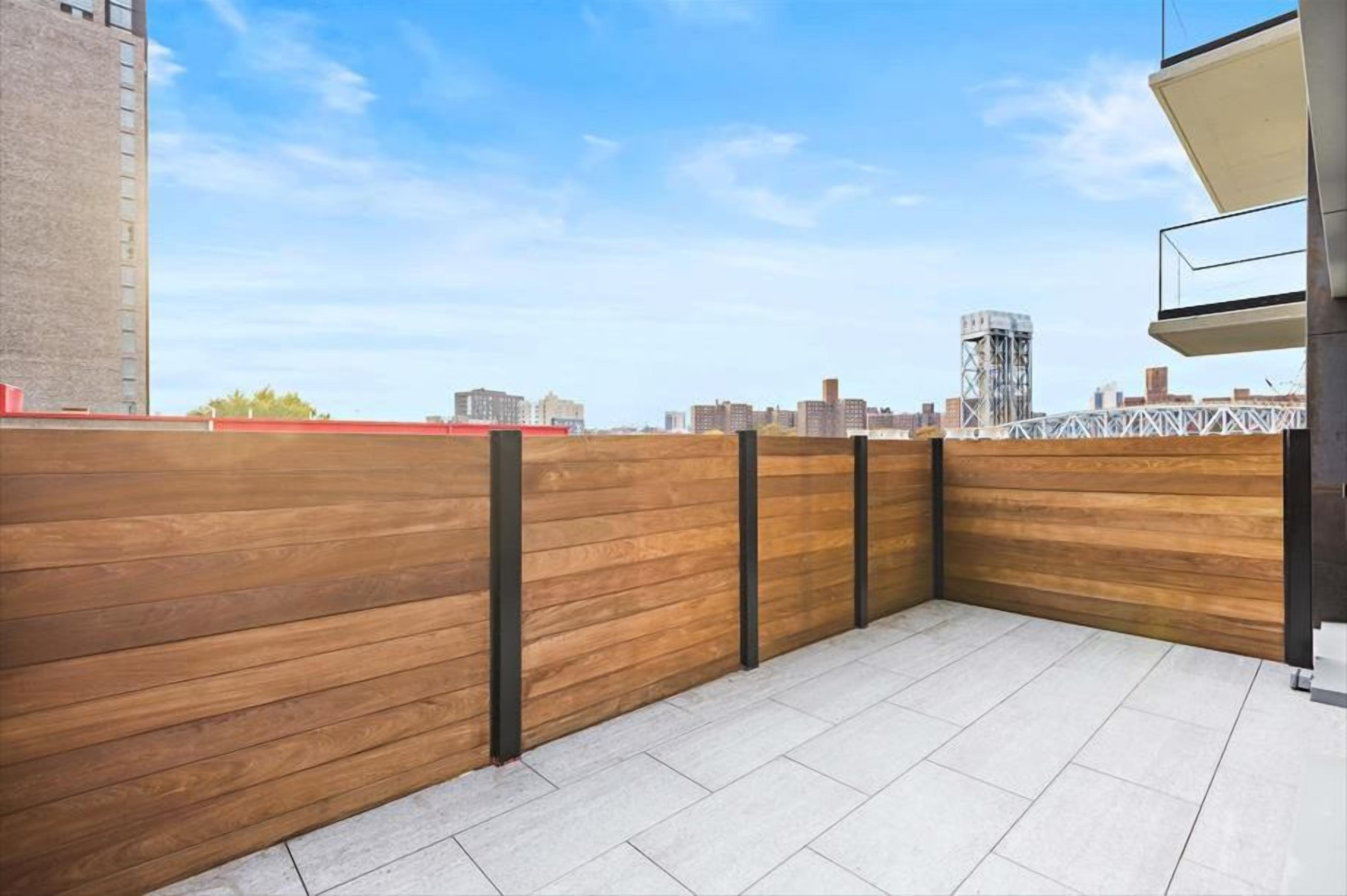 Een terras op het dak met een houten hek, betegelde vloer en een stadsachtergrond.