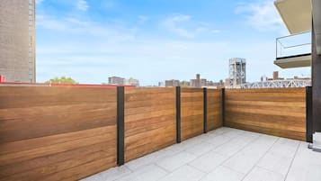 Een terras op het dak met een houten hek, betegelde vloer en een stadsachtergrond.