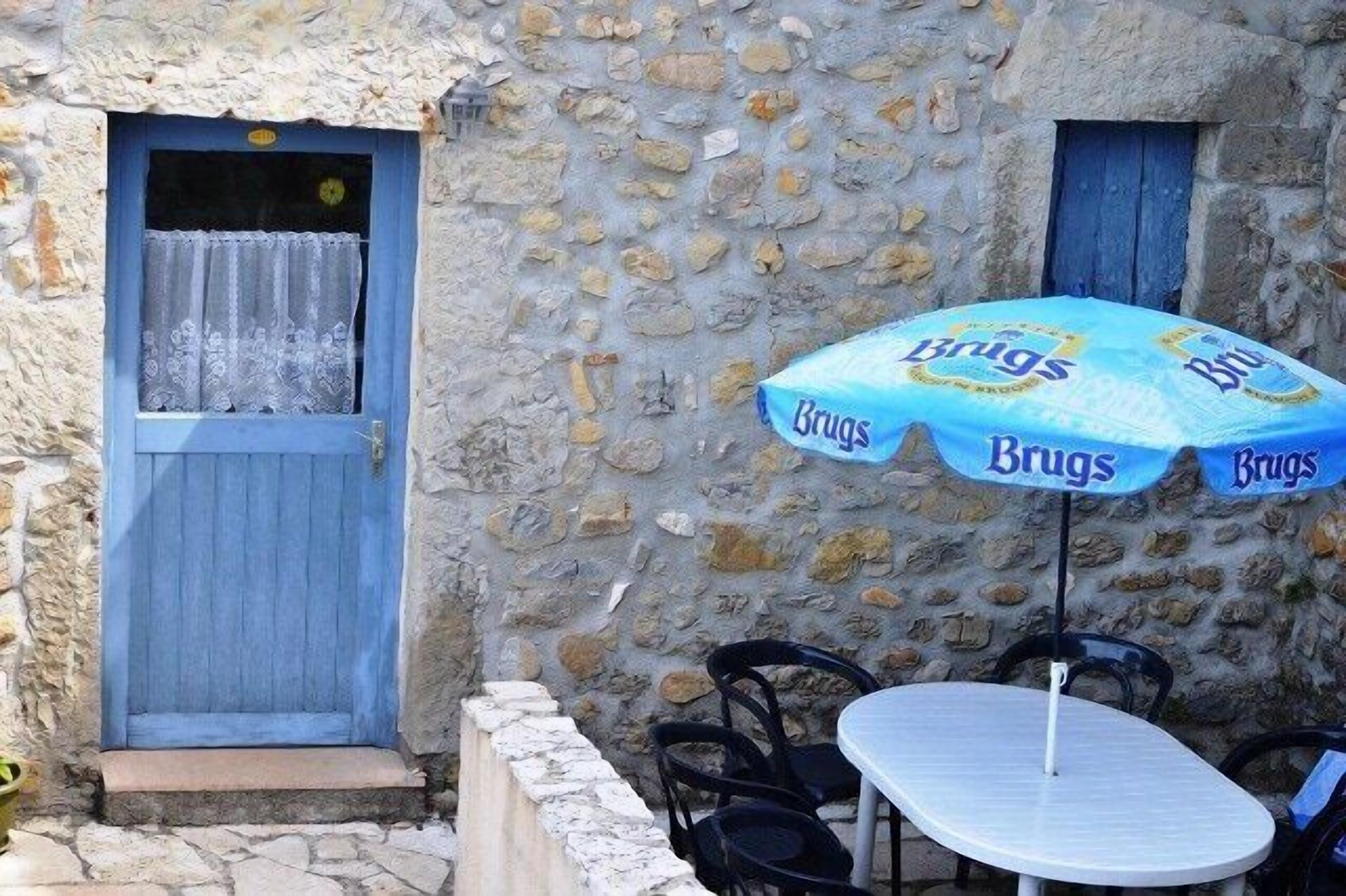 Een gebouw van steen met een blauwe deur en een raam, een tafel met stoelen en een blauwe parasol met de tekst 'Brugs'.
