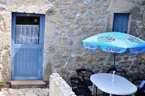 A stone building with a blue door and window, a table with chairs, and a blue umbrella with the word 'Brugs' on it. - Vacation Rental #11, Terrace & Pool, Domaine de Ribière (Grospierres)