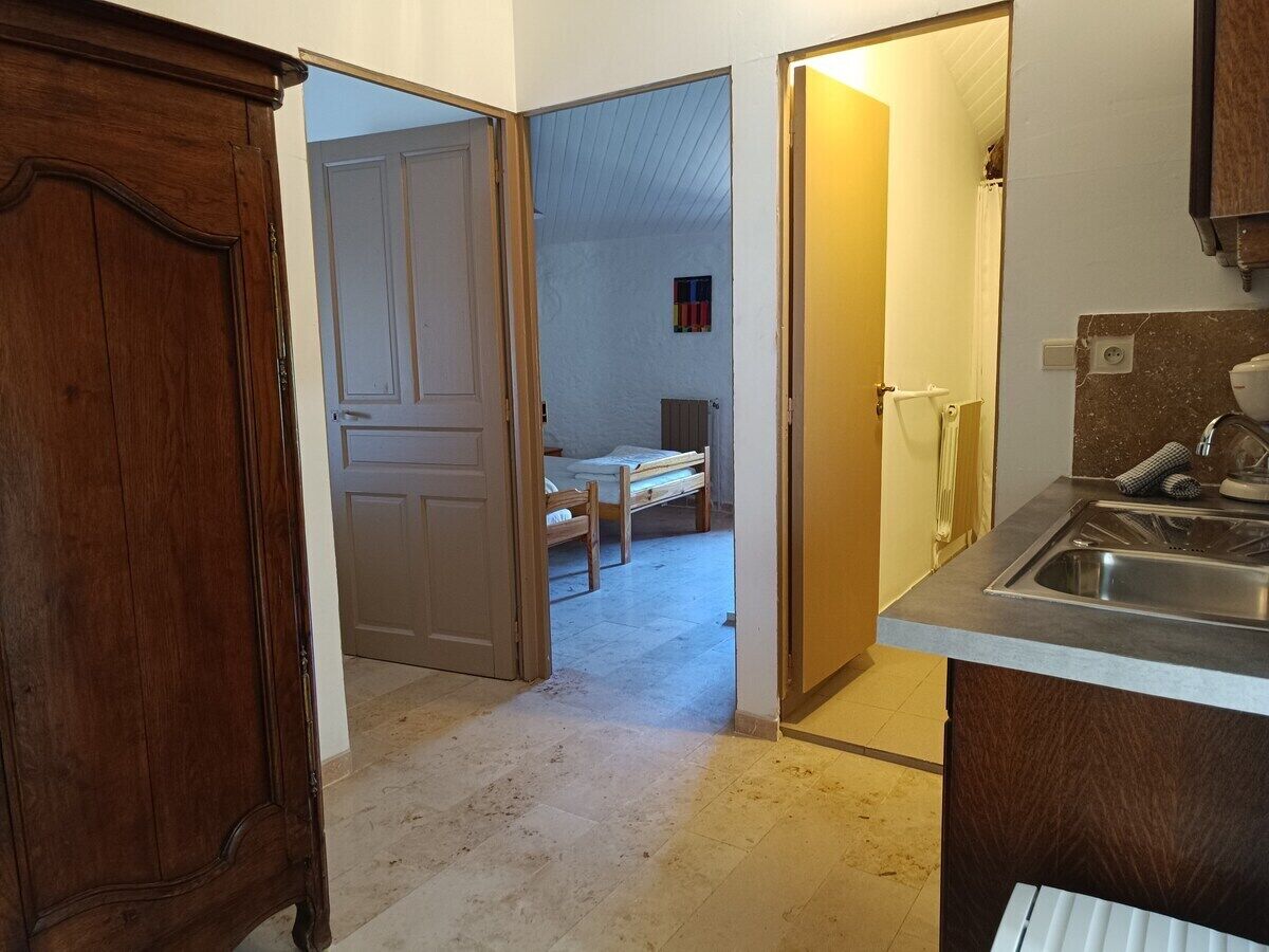 Une cuisine équipée d’un évier, d’un meuble en bois et d’une porte donnant sur une chambre à coucher avec un lit et une petite fenêtre.