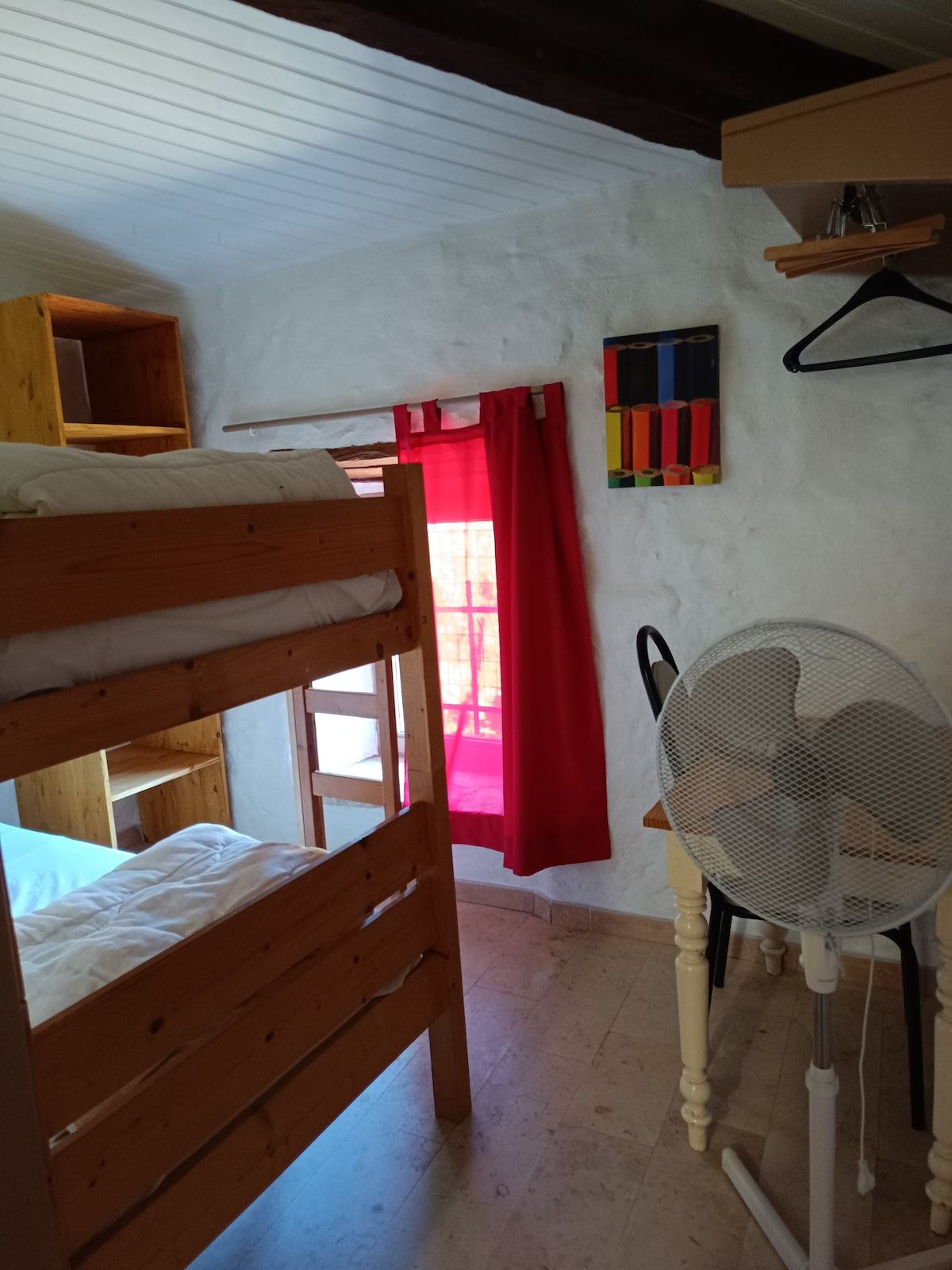 Ein Zimmer mit Etagenbett, Ventilator, rotem Vorhang und einem Regal mit diversen Gegenständen.