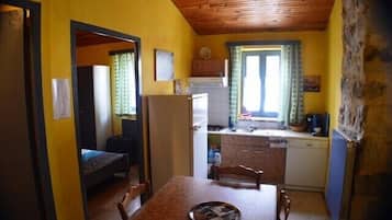 Een kleine keuken met een koelkast, spoelbak en raam. In de voorgrond staat een eettafel met stoelen.