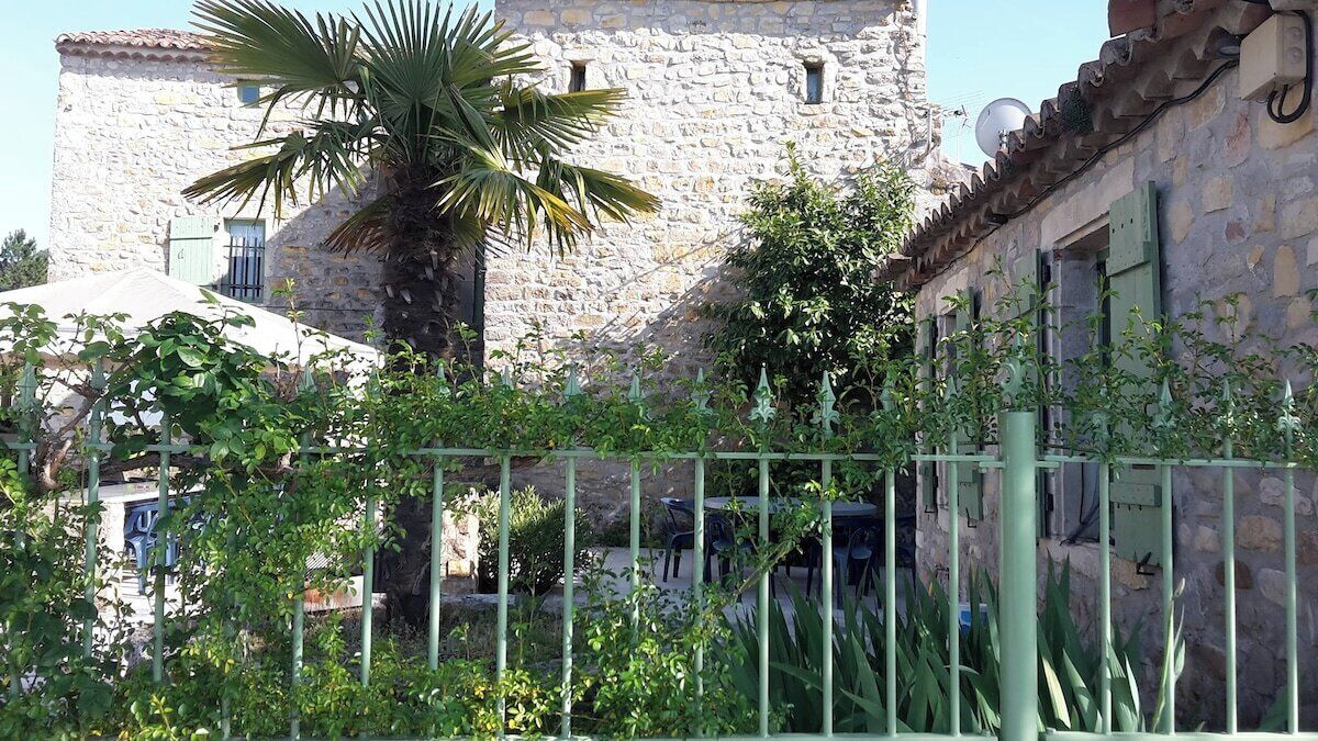 A stone building with a tower, surrounded by greenery and a metal fence.