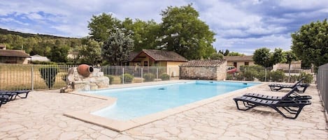 A swimming pool with stone tiles, lounge chairs, and a fenced area with a small building.