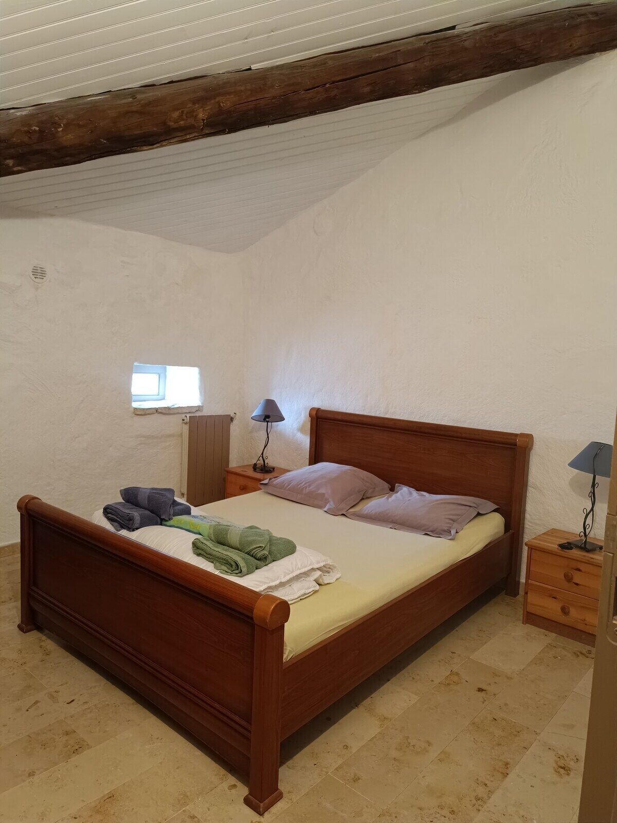 Een kamer met een enkel bed, een houten hoofdbord, een nachtkastje met lamp en een klein raam.