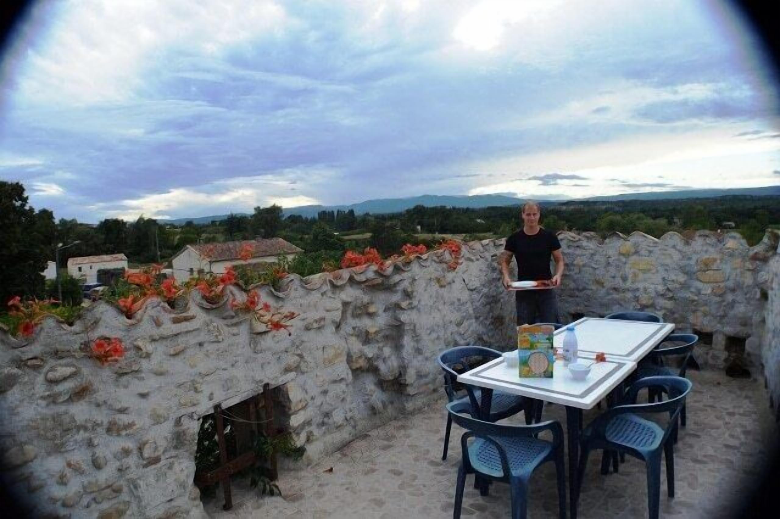 A table with chairs on a terrace with a stone wall, overlooking a landscape with houses and greenery.
