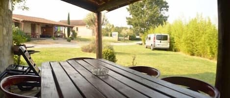 Una mesa de madera al aire libre con sillas, una vista despejada de un jardín con césped y una camioneta blanca al fondo.