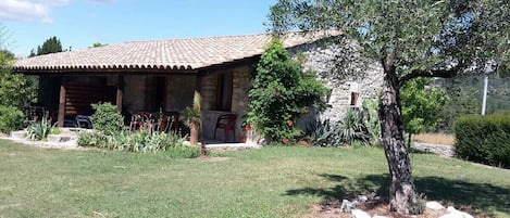 A rustic stone building with a tiled roof, a covered porch, and a well-maintained lawn with a tree and rocks.