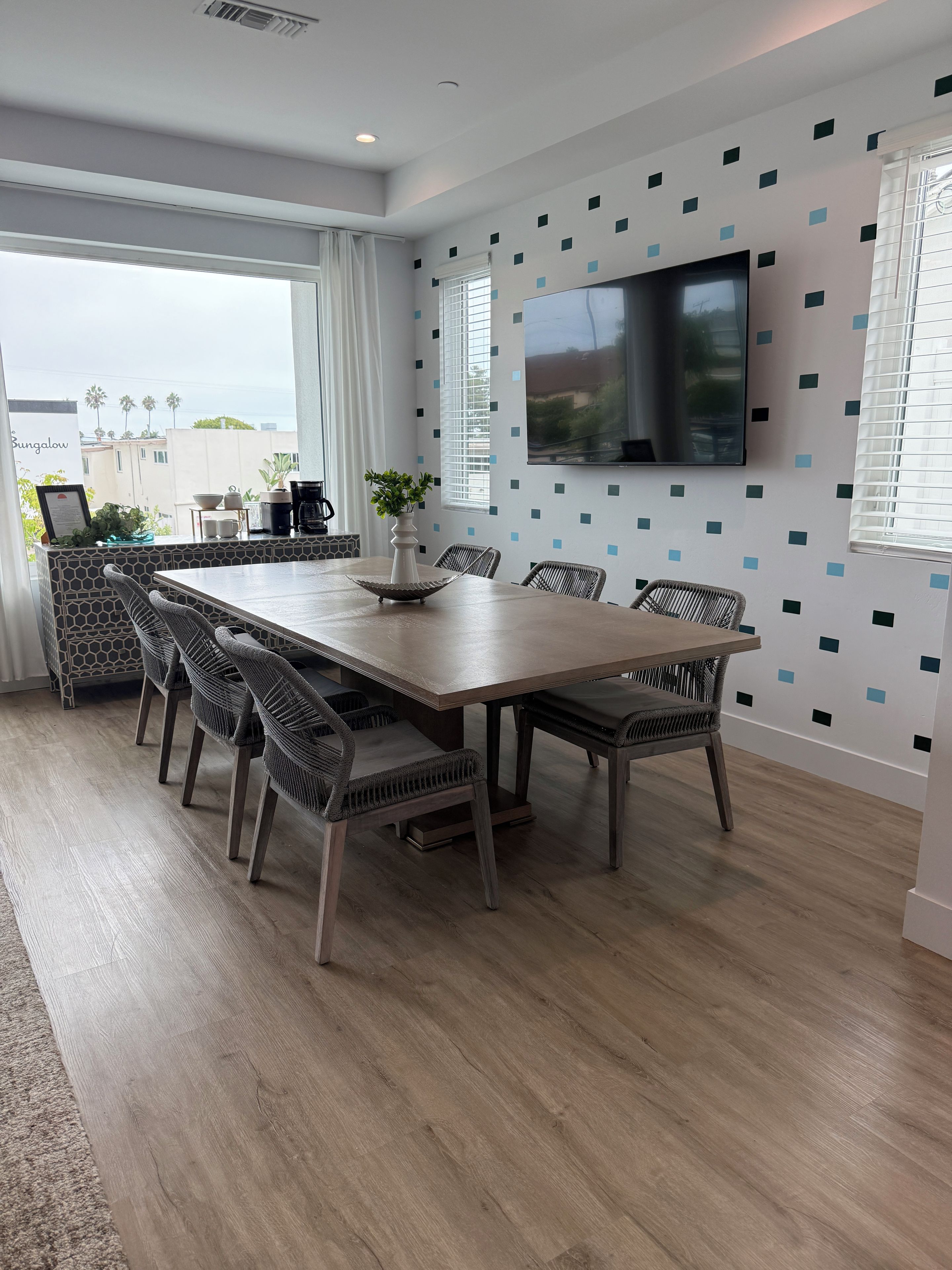 A modern dining area with a large table, wicker chairs, a potted plant, and a TV mounted on the wall.