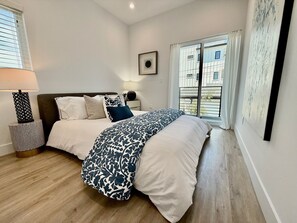 A bedroom with a bed, bedside table, lamp, and a window with curtains. - Beach Haus~Modern Comfort Spacious 4-bedroom in Pacific Beach/La Jolla (San Diego)